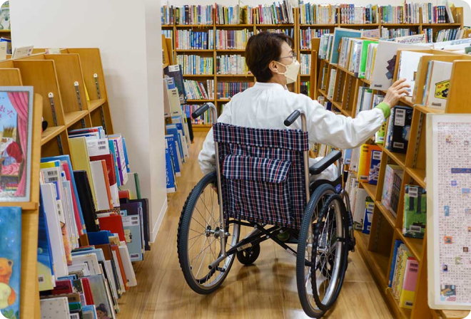 写真：図書館の本棚の間を車椅子で移動する女の人。通路がゆったりしているので、車椅子でも移動しやすいです。