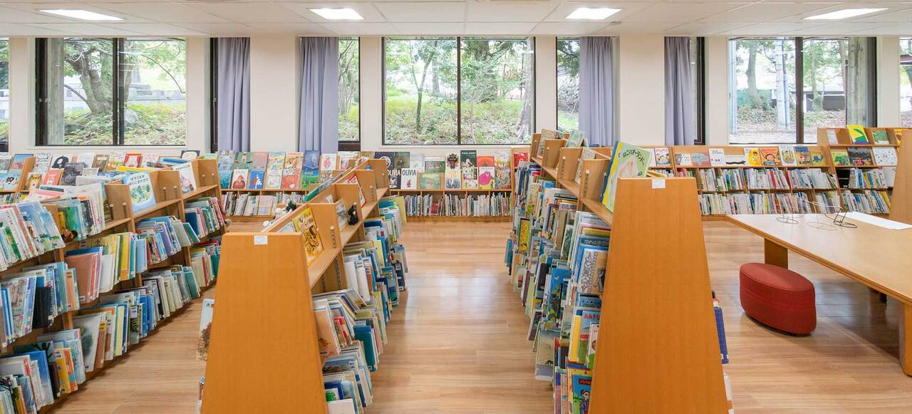 写真：図書館の中の様子。こどもが手に取りやすい高さの本棚が並んでいます。窓際の本棚にはたくさんの本の表紙が並んでいて、窓の向こうには緑が広がっています。